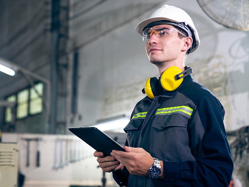 Engenheiro usando capacete de segurança e fones de ouvido, segurando um tablet enquanto observa atentamente algo fora da imagem. Ele está em um ambiente industrial iluminado.