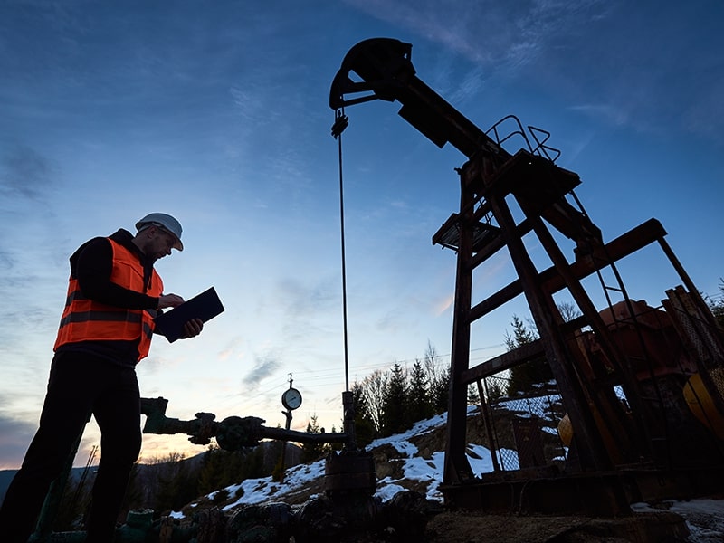 Silhueta de um engenheiro com capacete e colete refletivo, utilizando um tablet enquanto monitora um poço de petróleo ao amanhecer, com céu claro ao fundo.