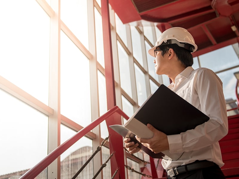 Engenheiro com capacete branco, segurando um bloco de notas, faz uma inspeção cuidadosa em um canteiro de obras. Ele está em uma escada vermelha, olhando atentamente para uma estrutura, enquanto avalia a integridade estrutural do edifício em construção ao fundo ensolarado.